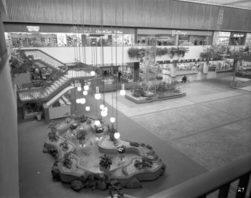 A spacious indoor mall with a central fountain, plants, and hanging lights, featuring shops on the upper level.