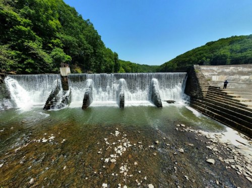 A serene waterfall cascading over a stone dam, surrounded by lush green trees under a clear blue sky.