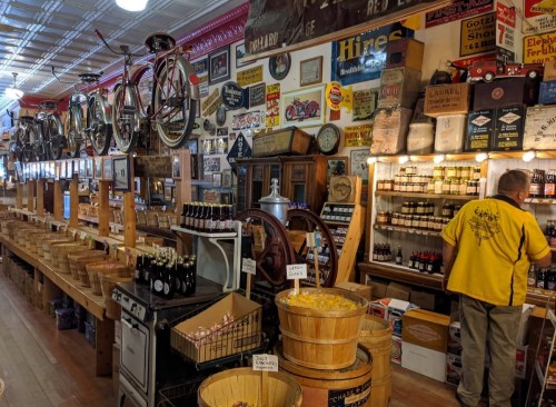 A vintage store interior filled with shelves of bottles, wooden barrels, and antique signs, with a person browsing.