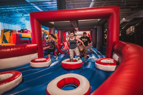 Children and adults play on a colorful inflatable obstacle course, jumping and running across the bouncy surface.