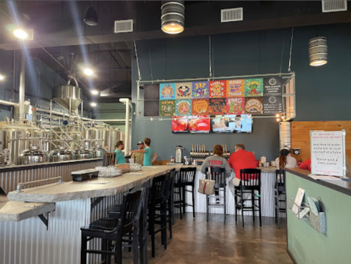 A modern brewery interior with a bar, patrons seated, and colorful beer menu displays on the wall.