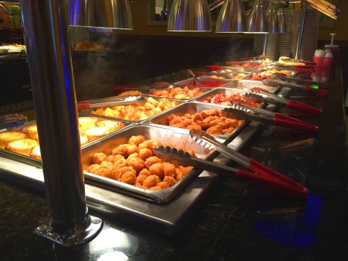 A buffet with various trays of food, including fried items, under warming lights, with tongs for serving.