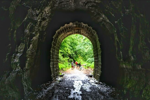 A view from inside a dark tunnel, leading to a bright, green landscape with people and bicycles outside.