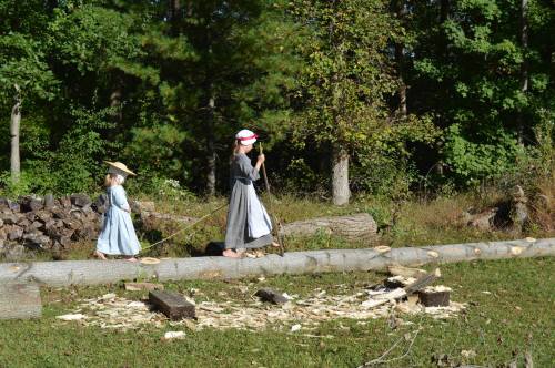 Two children in historical clothing walk along a log in a wooded area, surrounded by cut wood and greenery.