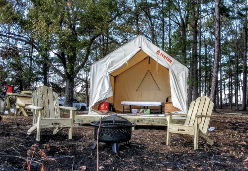 A cozy tent set up in a wooded area, with wooden chairs and a fire pit in front, surrounded by trees.