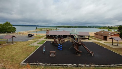 A playground with slides and climbing structures near a lake, surrounded by grassy areas and cloudy skies.