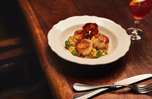 A plate of seared scallops on a bed of grains, garnished with greens, beside a cocktail on a wooden table.