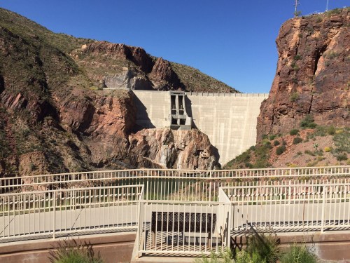 A concrete dam stands between rocky cliffs under a clear blue sky, with a railing in the foreground.