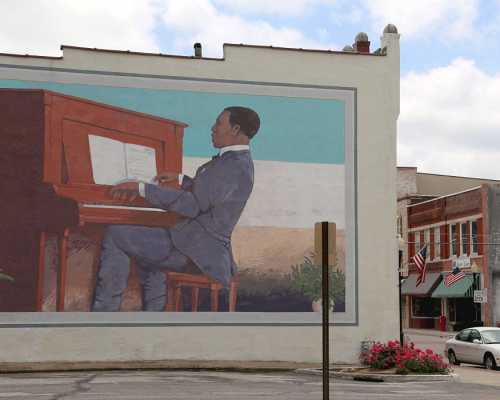 A mural of a man playing piano, seated at a wooden piano against a colorful background in a town setting.