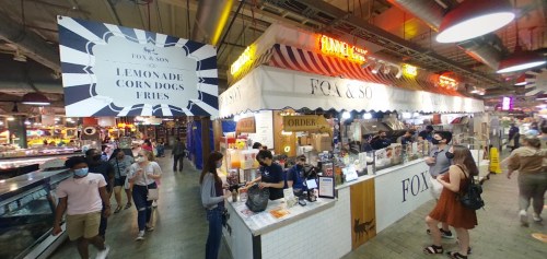 A bustling food stall named "Fox & So" offers lemonade and corn dogs in a lively market setting.