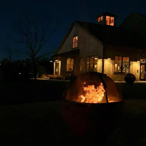 A cozy outdoor scene at night featuring a fire pit in front of a warmly lit barn-style building.