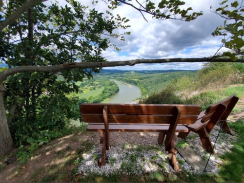 A wooden bench overlooks a winding river surrounded by lush green hills under a partly cloudy sky.