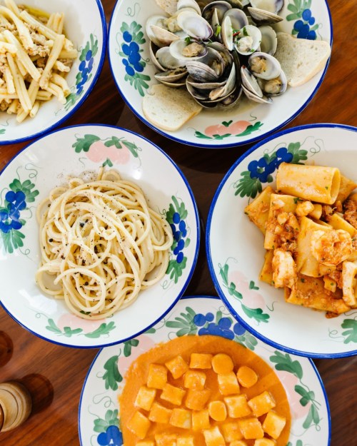 A colorful spread of pasta dishes, clams, and bread in decorative bowls on a wooden table.