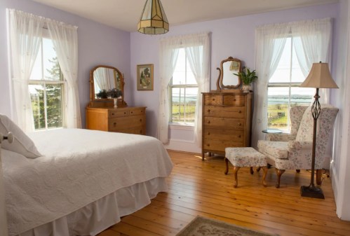 Bright bedroom with a white bedspread, wooden furniture, large windows, and a cozy armchair, overlooking a scenic view.