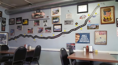 A diner wall decorated with a winding road mural, vintage car photos, and various memorabilia. Tables and chairs in foreground.