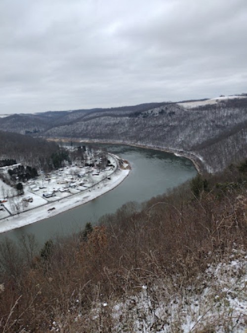 A snowy landscape with a winding river, surrounded by hills and trees, under a cloudy sky.
