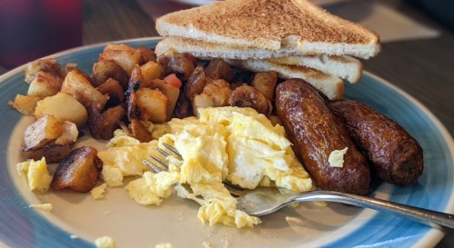 A plate of breakfast featuring scrambled eggs, sausage links, crispy potatoes, and toasted bread.