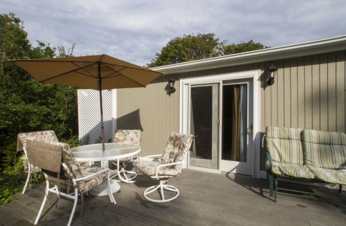 A patio with a round table, chairs, and an umbrella, next to a sliding door and surrounded by greenery.