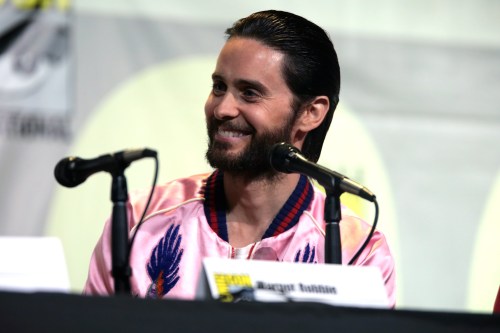 A smiling man with long hair wearing a pink jacket sits at a panel with microphones in front of him.