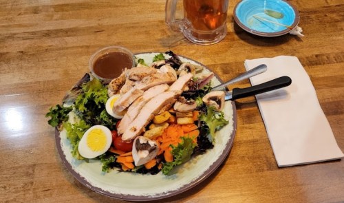 A colorful salad with grilled chicken, boiled egg, tomatoes, mushrooms, and a dressing on a wooden table.