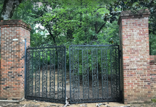 A wrought iron gate set between two brick pillars, leading into a lush, green area with trees.