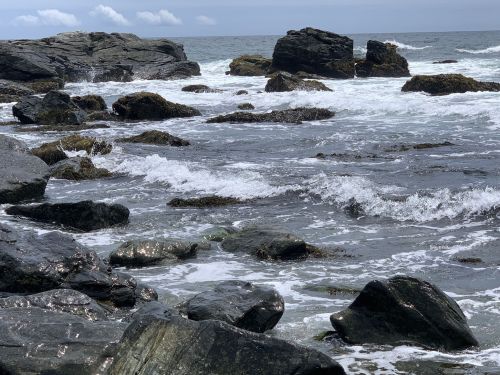 Rocky shoreline with waves crashing against boulders under a cloudy sky.