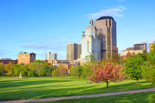 City skyline with modern buildings and a park featuring blooming trees under a clear blue sky.
