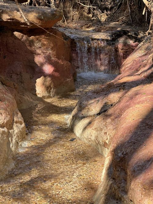 A small waterfall flows over reddish rocks into a shallow stream, surrounded by dry vegetation.