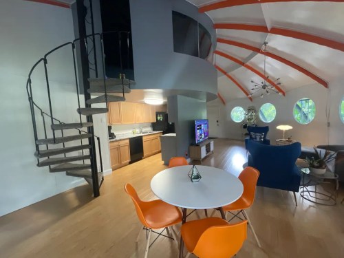 A modern living space featuring a spiral staircase, circular windows, and a dining area with orange chairs and a round table.