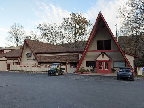A triangular-roofed lodge with a welcoming sign, surrounded by trees and parked vehicles in a scenic area.