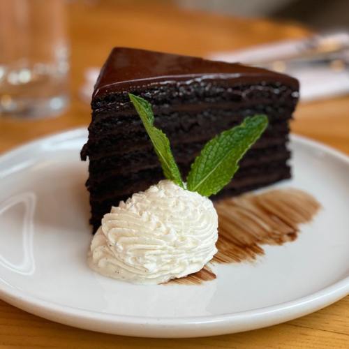 A slice of rich chocolate cake topped with glossy frosting, served with whipped cream and a mint leaf on a white plate.