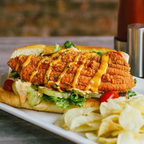 A crispy fried fish sandwich with lettuce, tomatoes, and sauce, served with a side of potato chips.