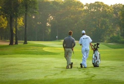 Two golfers walking on a lush green course, one carrying a golf bag, with trees and a flag in the background.