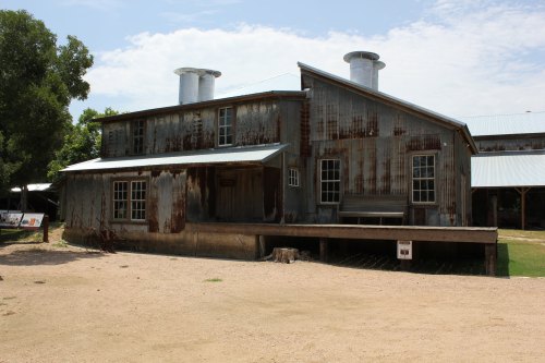 A weathered, two-story building with a metal exterior and two chimneys, set in a gravel area under a blue sky.