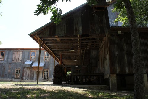 Rustic building with a corrugated metal roof, surrounded by trees and grass, featuring large windows and a weathered facade.