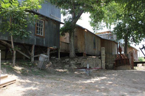 Rustic buildings on stilts surrounded by trees, with a picnic table and stone wall in a natural setting.
