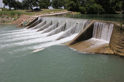 A series of small waterfalls cascading over a dam into a river, with kayakers visible nearby.