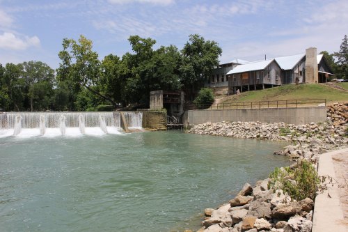 A serene river scene featuring a dam, flowing water, and a rustic building surrounded by trees and rocky banks.