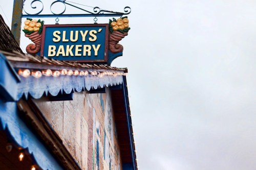 Sign for Sluy's Bakery hanging above a colorful building, with a cloudy sky in the background.