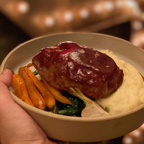 A hand holding a plate with meatloaf, mashed potatoes, sautéed greens, and roasted carrots.