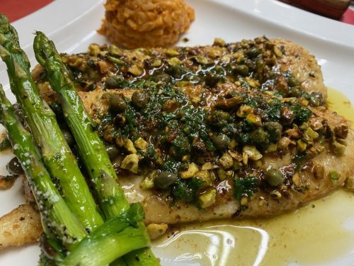 A plate featuring grilled fish topped with capers and herbs, served with asparagus and a side of rice.