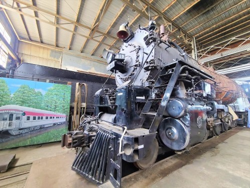 A vintage steam locomotive displayed indoors, with a mural of a train and trees in the background.