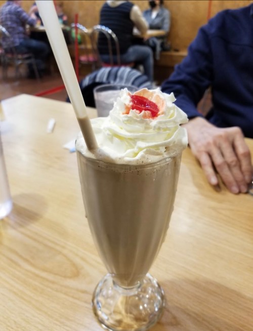 A tall glass of chocolate milkshake topped with whipped cream and a cherry, placed on a wooden table.