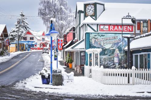 A snowy street scene featuring colorful buildings, a clock tower, and a sign for ramen in a quaint village.