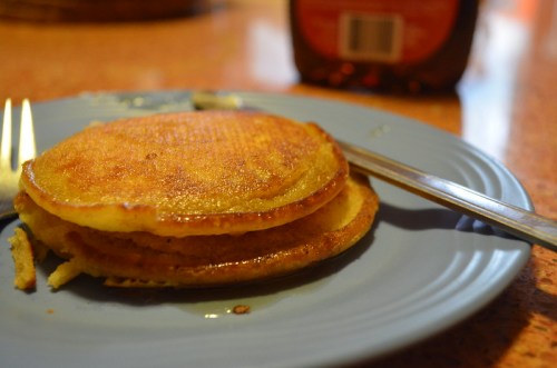 A stack of golden pancakes on a blue plate, with a fork beside them and a bottle of syrup in the background.