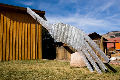 Giant metal fork sculpture leaning against a wooden building, set in a grassy area with a blue sky.