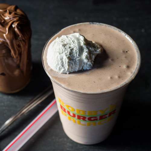 A thick milkshake topped with whipped cream, next to a jar of chocolate spread and a straw on a dark surface.