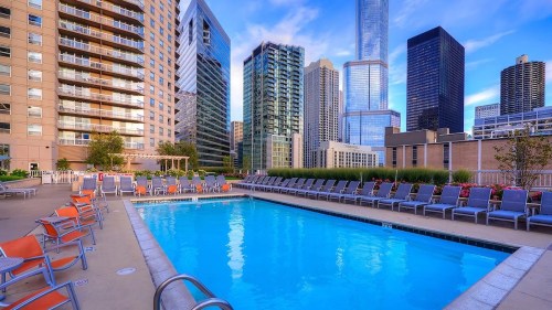 Rooftop pool surrounded by modern skyscrapers and lounge chairs under a clear blue sky.