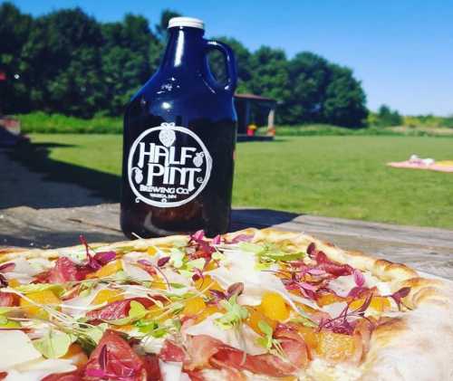 A close-up of a pizza topped with greens and meats, next to a growler from Half Pint Brewing Co. in a sunny outdoor setting.