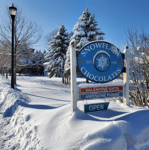 A snowy path leads to a sign for Snowflak Chocolates, advertising Valentine gifts and fudge, under a clear blue sky.
